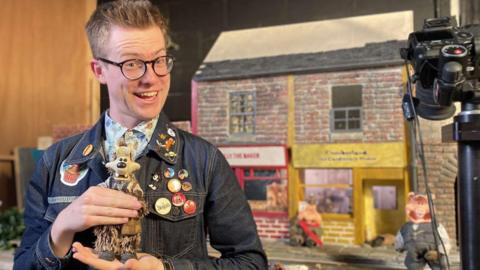 Man smiling while holding a clay wolf figure in front of a set depicting old-fashioned shops and the pigs from the story