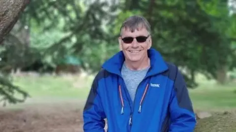 Steve Barber A man wearing sunglasses and a blue waterproof jacket smiles into the camera. He is standing outside under a large tree. More trees and an expanse of grass can be seen behind him.