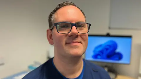 Nikki Fox/BBC Rob Bradley sitting in an urgent treatment centre. He is wearing dark blue scrubs and dark framed glasses. Behind him is a computer screen. 