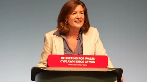 Eluned Morgan smiling as she gives her speech to the Welsh Labour conference. She is standing behind a podium with red sign with the words "Delivering for Wales - Welsh Labour" on it in white lettering. She is wearing a light jacket and grey blouse