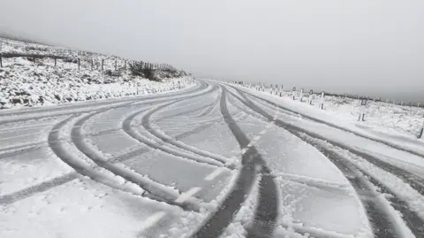 DOI Snowy conditions on the Mountain Road at the Hailwood's Rise. Fence posts and road markers line the snow covered road which has the tyre tracks of vehicles on it. There is snow cover on the the grass at either side.