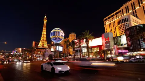 Reuters A white sportscar travels down the Las Vegas strip. The Eiffel Tower replica can be seen lit up, along with a hot air balloon type structure and large cream buildings, with palm trees lining the road. 