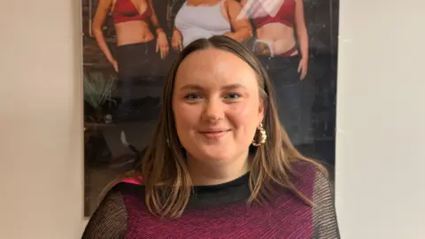 Ellie Brown with long brown hair and gold earrings wearing a black see through top with a hot pink vest underneath. She stands under a poster with three models wearing Recondition clothing. She is smiling.