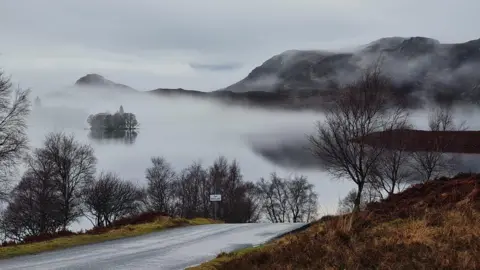 Laura Kennedy A lake is hidden by fog. A road emerges out of the fog with bare trees on each side. Hills are seen in the background and it's a cloudy day. 