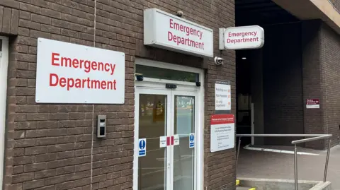 BBC A picture of the front doors of the Emergency Department in St Helier, Jersey. The hospital building is made up of brown bricks, and the doors are glass.