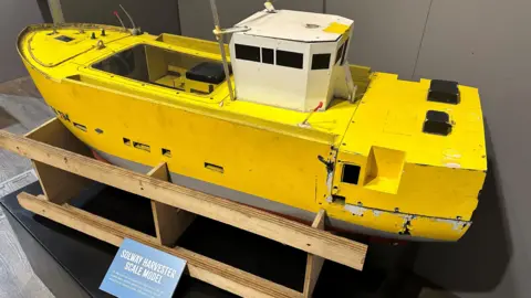 A full scale model of he scallop dredger, which is yellow with a white wheelhouse, placed on a wooden stand with a blue information board in front of it.