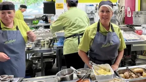 Dinnertime at the Whale Hill Primary School kitchen in Eston. Lunch staff in dark hats, lime green tops and grey aprons standing in front of silver hot plates of food including baked potatoes, baked beans, crusty bread, carrots and cucumber.