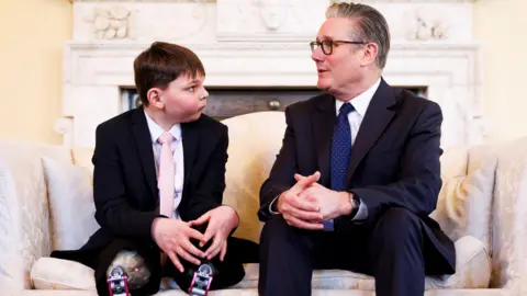 PA Media A boy, wearing a black suit and pink tie, sat next to a man wearing a black suit and blue tie.