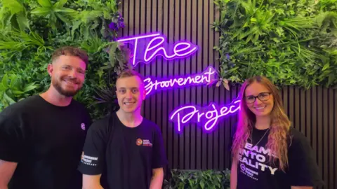 A picture of two men and a woman in front of a purple neon sign. The sign reads "The Groovement Project".  They are all wearing black t-shirts.