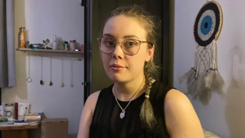 A blonde woman wearing glasses who has her hair in a side plait and is wearing clear circular glasses and a black racer top. She is also wearing a sliver love heart necklace.