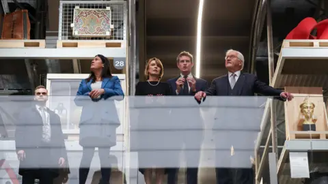 Reuters Steinmeier, his wife, Lisa Nandy and Tristram Hunt stand on a balcony in the V&A Storehouse. They are stood in a line leaning against the glass balcony.