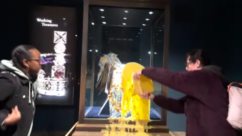 Take Back Power A protester pours custard on the display case containing the Imperial State Crown in the Jewel House at the Tower of London while another protester watches on.