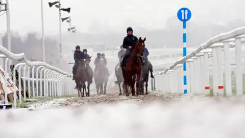 PA Media Several jockeys are exercising their horses on a snow covered track at the Cheltenham Festival on the second day of the four day event. They are trotting between some white barriers.