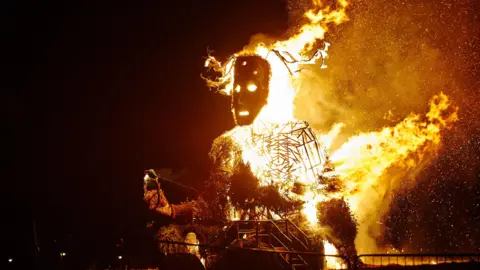 A giant Green Man sculpture has been lit on fire in the dark. Its face is illuminated with large flames coming from its torso. 