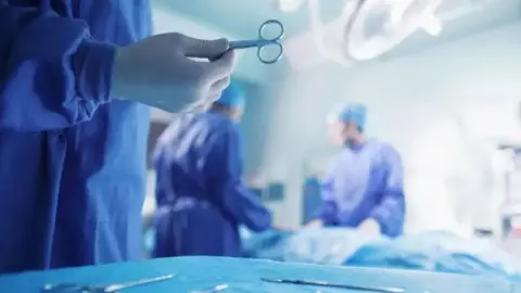 A generic picture shows three surgeons in blue scrubs one of whom is holding a pair of scissors