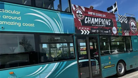The bus parked before the players board it. There is a St Piran flag flying on the top deck and a banner that reads: WE'RE GOING UP! CHAMPIONS