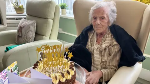 A woman is seated in a high‑backed, light‑coloured armchair upholstered in what appears to be leather or faux leather. The person is positioned slightly to the right side of the frame and faces the camera directly. She has short, light grey hair and is wearing a light‑coloured, buttoned shirt with narrow vertical stripes. A dark, soft-looking garment or blanket is draped around the shoulders and upper arms. The person’s hands rest near the lap or armrest area. Directly in front of the seated person, resting on a low surface or table, is a round cake placed on a clear plastic tray.
The cake is decorated with piped cream around the top edge. Several curved chocolate pieces and small decorative elements are arranged evenly along the top. A gold decorative topper is placed in the centre of the cake. The topper contains readable text that says “Happy 100th Birthday”, with star shapes incorporated into the design.