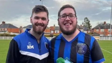 Harry McCabe Harry McCabe and his brother Jack. They both have short brown hair and beards. Jack is wearing glasses and a blue and black football shirt while Harry is wearing a blue, white and black sports top. They are stood on a football pitch with houses in the background.
