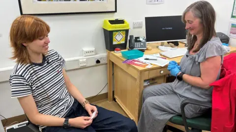 BBC/Mark Ansell A young woman at a vaccination appointment in a pharmacy consultation room. The patient sits opposite a healthcare worker wearing blue gloves, with information leaflets and medical equipment visible on a desk. The young woman has short, reddish-brown hair and is wearing a navy and white striped short-sleeved T-shirt, dark blue jeans, a black wristwatch and a beaded bracelet. The person seated on the right has shoulder-length grey hair and is wearing a light grey blouse with ruffled short sleeves and matching grey trousers. 