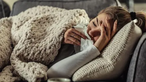 Woman lies on a sofa looking unwell, with a knitted blanket pulled over herself. She holds a tissue in one hand and clutches her forehead, her eyes are closed.