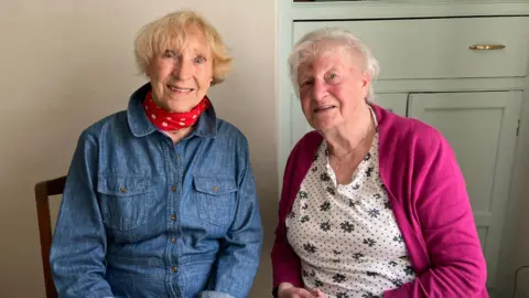 Two women sit next to each other and look directly at the camera. The woman on the left is wearing a denim shirt and a red, dotted scarf, while the woman on the right is wearing a floral top and magenta cardigan.