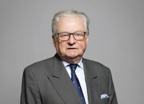 An official House of Lords photograph of Lord Elis-Thomas. He is looking directly at the camera and wearing glasses, a grey suit, light blue shirt and dark blue tie.  