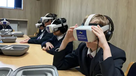 A few children with grey VR headsets on. They have a pair of black headphones on as well a dark navy blue school uniform.