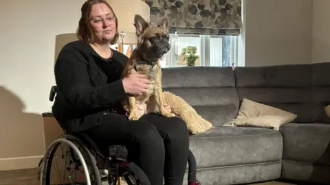 Matt Precey Young woman in a wheelchair playing with a dog. The dog is brown coloured. The woman has brown hair and is wearing glasses. She is in a living room. Behind her, there is a grey sofa and a window with a brown-patterned blind. 