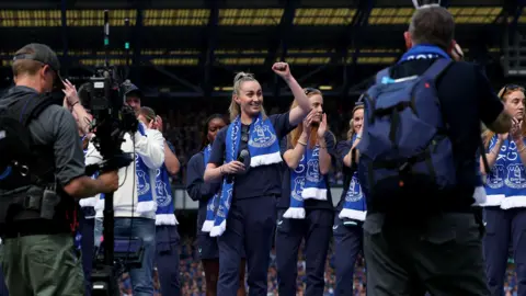 Members of Everton's women's team clap while standing on a podium on the Goodison Park pitch during a ceremony to mark the men's last game at the ground. Howard Kendall Gwladys Street End can be seen in the background.