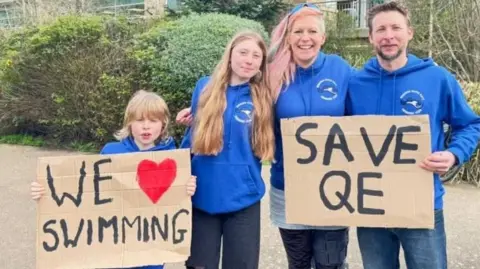 Elizabeth Mills with her children and partner holding signs saying "we love swimming" and "save QE"
