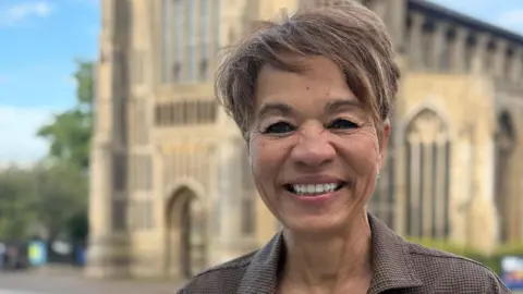 Tessa McWatt standing outside The Forum in Norwich in front of a church which is out of focus. She is looking directly at the camera and is smiling. She has short brown hair.
