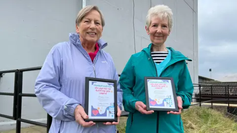 BBC Two women stand together next to a large grey building holding certificates. On the left, Angela Turner has short grey hair and is wearing a purple coat. On the right, Joan Woodhouse has short white hair, and is wearing a green coat.