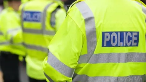A stock image of police officers wearing yellow high-viz police jackets. The back of an officer's jacket, with the word police is in the foreground. The background is blurred, with other officers, who are all unidentifiable, standing together.