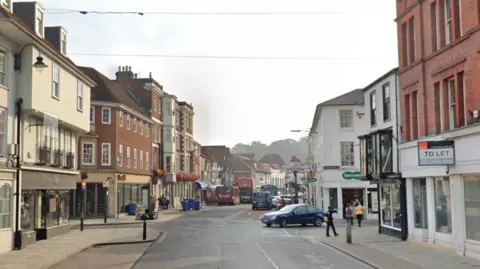 Google maps view of New Canal and Milford Street in Salisbury, Wiltshire. Shops can be seen lining the street and vehicles can be seen in the distance on the road. A few pedestrians can also be seen walking on the pavement.