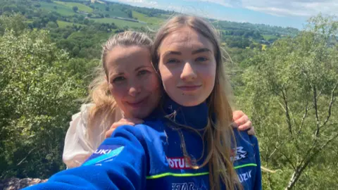 Family Handout Leanne and Lily are stood on a hill taking a selfie with hills and trees behind them. They are stood close together smiling. Lily has straight dark hair and Leanne's is pulled back
