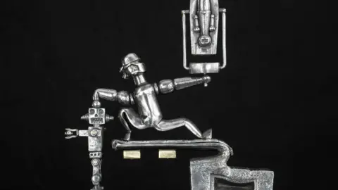 Cooper Gallery/Sam Sherborne A silver metal sculpture depicts a human figure straddling a pedestal, holding an invisible statue in his right hand and punching a robot with his left hand.