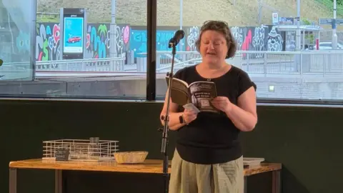 Penny Blackburn A woman is standing at a microphone and reading from "Words on a NE street". The views out of the Baltic building can be seen through the window behind her, including a bus stop and graffiti artwork.