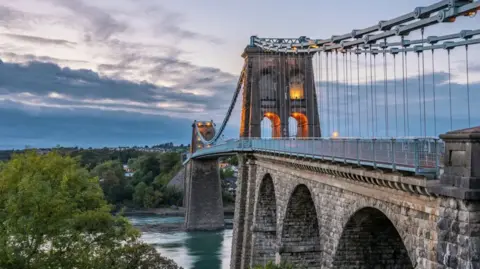 The Menai Bridge is made of stone, with blue water flowing beneath. Green trees to the left hand side. A cloud sunset in the background.