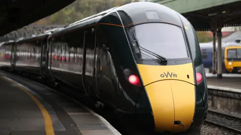 A picture of a GWR train parked at a train station. The body of the train is green with the front a yellow stripe.