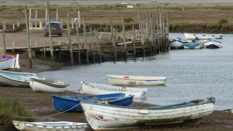 White and blue fishing boats at Morston Quay by the water. An access road and wooden jetty are at the edge of part of the water. 