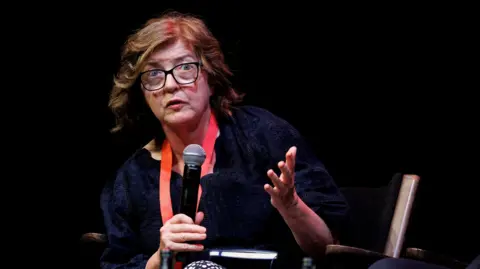 Baroness Sue Gray sitting on a chair holding a microphone. She wears a red lanyard and has short brown hair and glasses. 