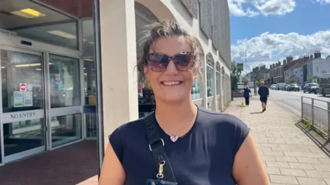 Andrew Turner/BBC Sadie Laurence is wearing sunglasses and a black top, with a bag strap over her right shoulder. She has long dark hair, tied back. The building is a 1970s design, featuring brown bricks, concrete pillars and arches, several infilled with aluminium-framed windows and windows, and flint-faced concrete panels on the upper storey.