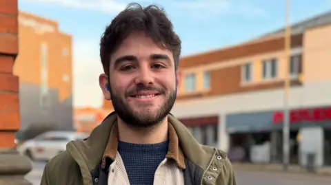 Jake Holland, who has dark hair and a beard, is smiling and looking directly at the camera. He is wearing a khaki jacket, cream shirt and grey jumper.