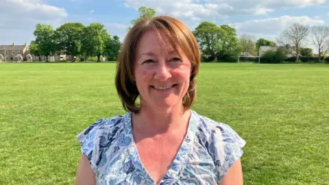 Vikki Irwin/BBC Dr Catherine Judkins smiles at the camera while standing in a sports field. She has red hair that has been cut in a bob. She wears a blue and white blouse.