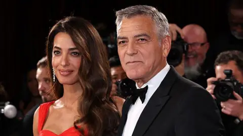Amal Clooney is dressed in a red dress while husband George can be seen in a tuxedo and black bow tie next to her. A bank of photographers are in the background out of focus