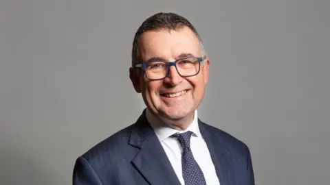 UK Parliament Sir Bernard Jenkin, who is wearing a suit, tie and glasses. He is smiling at the camera