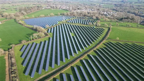A drone shot on a clear day showing solar panels stretching across a large area of fields, with blocks of various shapes bisected by hedgerows and then green fields surrounding the development with wooded areas and the village with its white stucco homes and brown roofs, some close to one end of the solar park, but with a significant wooded area in between . 