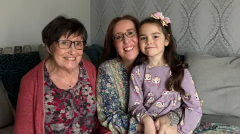 Two woman and a child in the picture. The woman on the left is wearing a red cardigan, a patterned top, has short dark hair and is wearing glasses, she is smiling at the camera. Next to her is a woman in a floral long sleeve top, wearing glasses with shoulder length brown hair and she is smiling. On her knee is a little girl with a long sleeved purple top and long dark hair. They are all sat on a grey sofa.