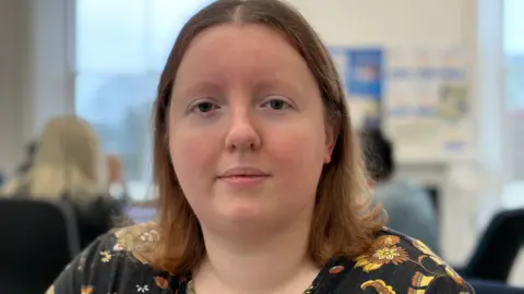 BBC Woman with shoulder length light brown hair and wearing a black top with flowers on. 