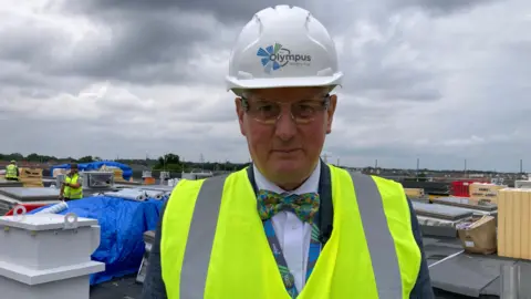 A man wearing a grey suit and colourful bow tie is looking at the camera. He is wearing a yellow high vis jacket over his suit and a hard hat, which reads 'Olympus Academy Trust'. He is standing on the roof of a building where construction is taking place.